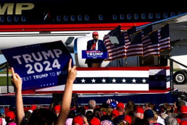 man wearing suit and red hat stands at podium in front of crowd of people holding signs and wearing red hats